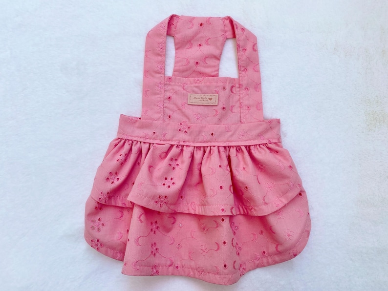 May include: A pink dog dress with eyelet detailing and a ruffled skirt. The dress has adjustable straps and a small tag near the top. The fabric has a floral pattern and is made of a lightweight material.