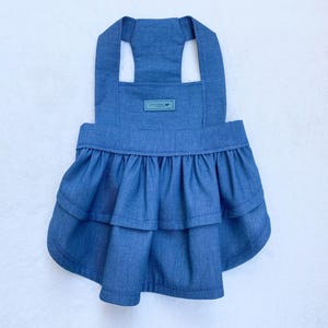 May include: A denim blue dog dress with a layered ruffle skirt and halter straps. A small rectangular tag is sewn on the front. The dress is designed for a small dog and is made of a soft, lightweight fabric.