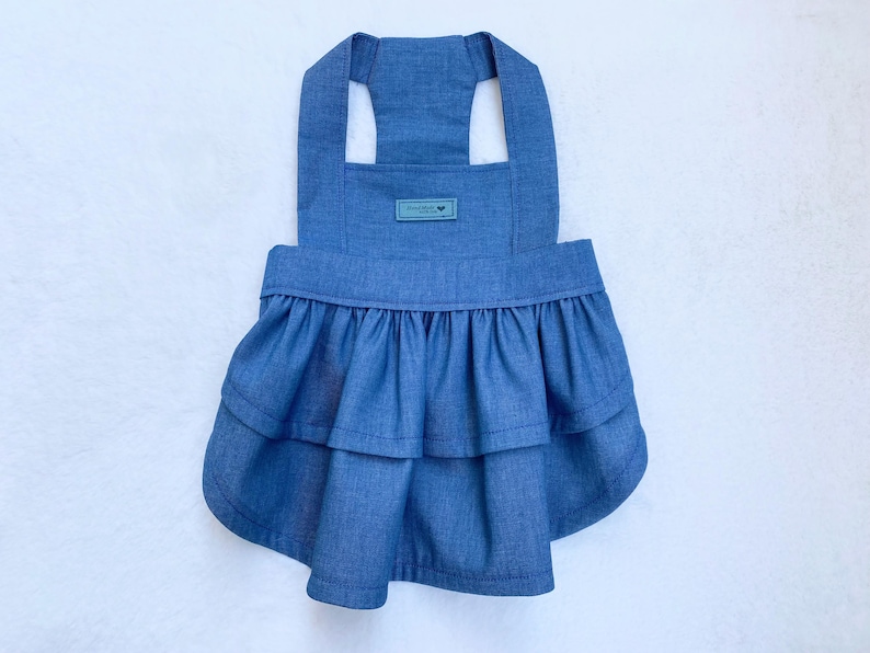 May include: A denim blue dog dress with a layered ruffle skirt and halter straps. A small rectangular tag is sewn on the front. The dress is designed for a small dog and is made of a soft, lightweight fabric.