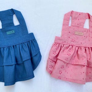 May include: Two pet dresses. One is denim blue with a frilled skirt and a small rectangular tag. The other is pink with a floral eyelet pattern and a frilled skirt. Both have halter-style straps.