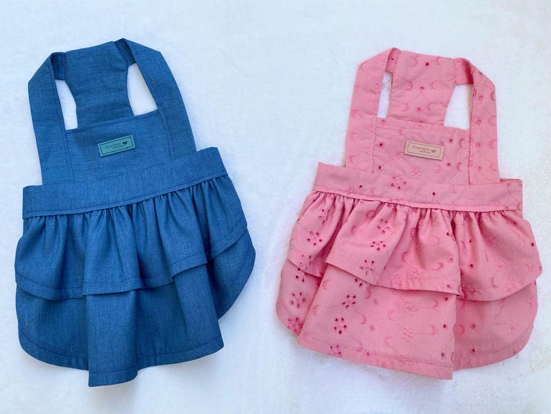 May include: Two pet dresses. One is denim blue with a frilled skirt and a small rectangular tag. The other is pink with a floral eyelet pattern and a frilled skirt. Both have halter-style straps.