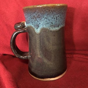 May include: A handmade ceramic mug with a unique design. The mug features a dark brown base with a blue and brown top. The handle has a distinctive thumb rest. The mug is approximately 13 cm tall.