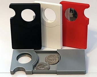 May include: A gray coin purse with a hinged lid that opens to reveal two coin slots. The purse is closed and sitting on a white surface next to three other coin purses. The other purses are black, white, and red.