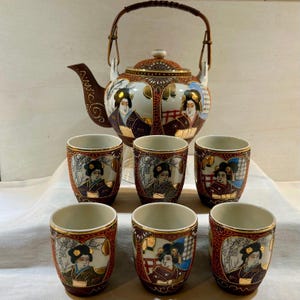 May include: A ceramic tea set featuring a teapot with a brown handle and six matching cups. Each piece is decorated with gold-trimmed illustrations of figures in traditional attire against a dark red and white background with intricate patterns.