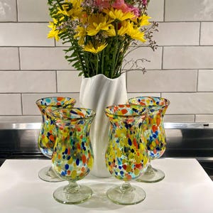 May include: Four colorful, speckled glass goblets with a clear stem and base. The goblets are filled with yellow, red, blue, and green spots. A white vase with a bouquet of yellow and pink flowers sits behind the goblets.