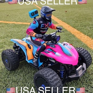 May include: A remote-controlled four-wheeled all-terrain vehicle with a bright pink and blue body, featuring a rider figure wearing a black, blue, and orange FXR branded racing suit and helmet, with a blue action camera mounted on the back.