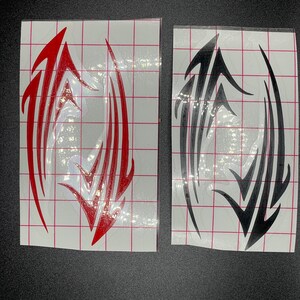 Pu&ograve; includere: Due adesivi in ​​vinile, uno rosso e uno nero, con un design in stile tribale. Gli adesivi sono su uno sfondo bianco a griglia.