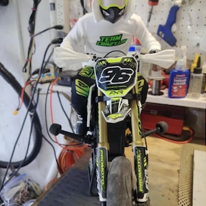 May include: A miniature motorcycle with gold and black accents, featuring the number 96 on the front. The rider is wearing a white hoodie with "TEAM CRAZY" printed on it, and a helmet. The motorcycle has "PRO MOTO" branding.