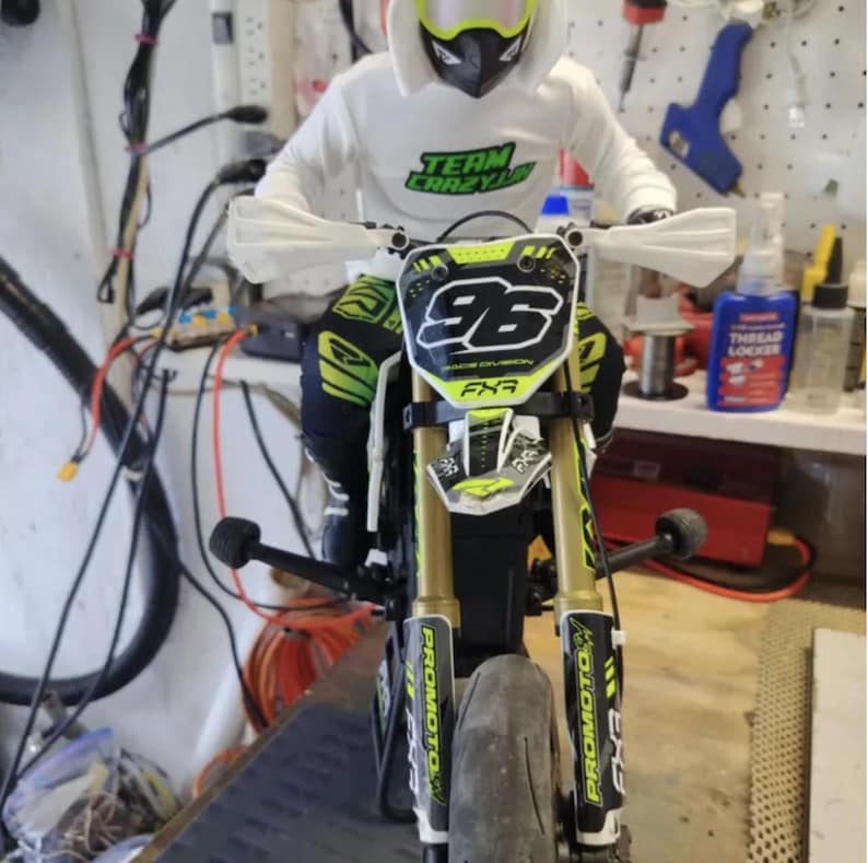 May include: A miniature motorcycle with gold and black accents, featuring the number 96 on the front. The rider is wearing a white hoodie with "TEAM CRAZY" printed on it, and a helmet. The motorcycle has "PRO MOTO" branding.