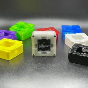 May include: A white, 3D printed box with the text "GNSS Performance Analyzer GSM020" on the front. The box is surrounded by other 3D printed boxes in various colors, including purple, green, yellow, red, blue, white, and black.