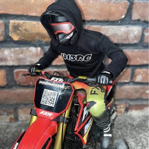 May include: A miniature red and black dirt bike with gold forks, a black rider wearing a black hoodie with "L3RC" printed on it, and a helmet with red-tinted goggles. The bike has "FXR" and "PROMOTOMX" branding.