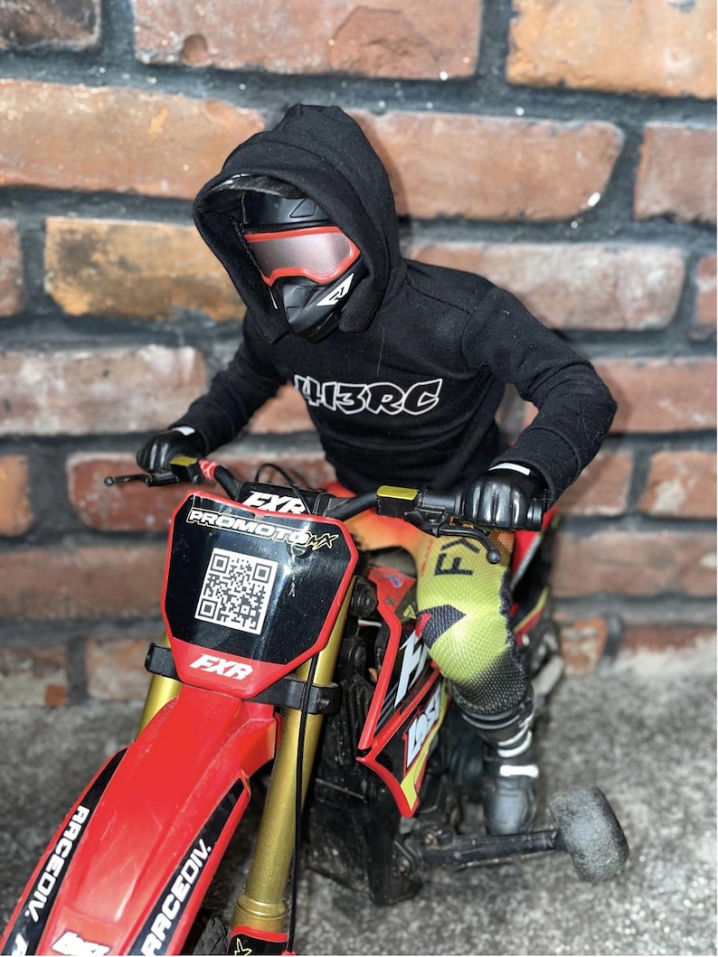 May include: A miniature red and black dirt bike with gold forks, a black rider wearing a black hoodie with "L3RC" printed on it, and a helmet with red-tinted goggles. The bike has "FXR" and "PROMOTOMX" branding.