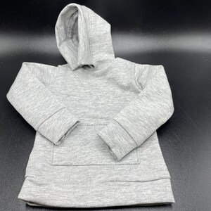 May include: A small, light gray hoodie with a hood and a kangaroo pocket. The miniature sweatshirt is made of a soft, textured fabric. The sleeves have ribbed cuffs, and the bottom hem is also ribbed. The hoodie is designed for a child or a doll.