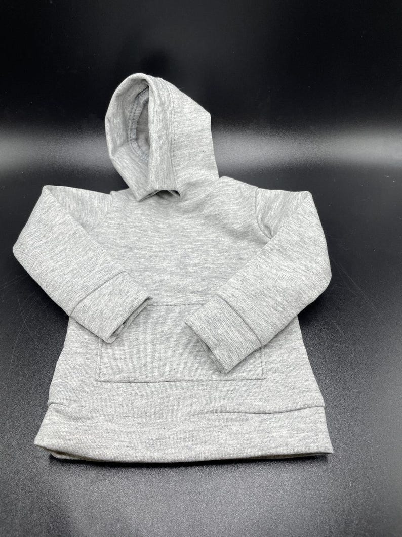 May include: A small, light gray hoodie with a hood and a kangaroo pocket. The miniature sweatshirt is made of a soft, textured fabric. The sleeves have ribbed cuffs, and the bottom hem is also ribbed. The hoodie is designed for a child or a doll.