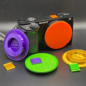 May include: A black Sony Alpha camera with a purple, orange, and green lens cap. The lens cap is made of plastic and has a textured surface. The camera is sitting on a black surface.