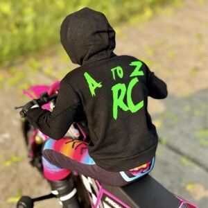 May include: A black hooded sweatshirt with the text "A to Z RC" in bright green on the back. The person is sitting on a pink and white toy motorcycle, wearing colorful leggings. The background is blurred.