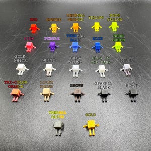 May include: A collection of small, robot-shaped figurines in various colors, including red, orange, yellow, pink, purple, blue, green, white, grey, silver, brown, black, and gold. Each figurine has a different color name printed above it.