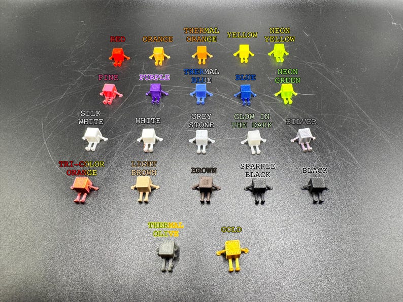 May include: A collection of small, robot-shaped figurines in various colors, including red, orange, yellow, pink, purple, blue, green, white, grey, silver, brown, black, and gold. Each figurine has a different color name printed above it.