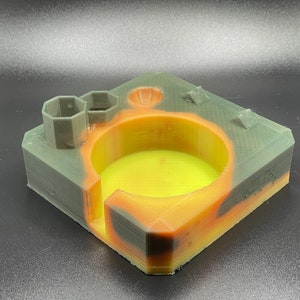 May include: A 3D printed multi-colored cup holder with a hexagonal compartment and a round cup holder. The cup holder is printed in a camouflage pattern with green, brown, and orange colors.