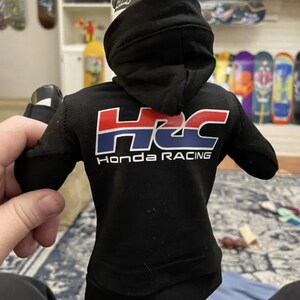May include: Black hooded sweatshirt with the Honda Racing logo in red, white, and blue. The sweatshirt has a drawstring hood and long sleeves. The text "Honda Racing" is printed below the logo.