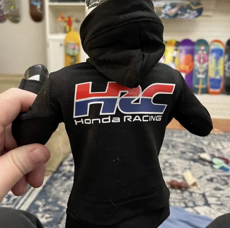 May include: Black hooded sweatshirt with the Honda Racing logo in red, white, and blue. The sweatshirt has a drawstring hood and long sleeves. The text "Honda Racing" is printed below the logo.