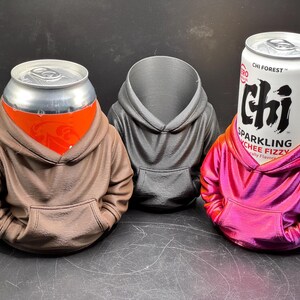 May include: Three hoodie-shaped can holders in brown, gray, and pink. The brown holder has a can with an orange label, the gray holder is empty, and the pink holder has a can with the text "CHI FOREST" and "SPARKLING LYCHEE FIZZY".