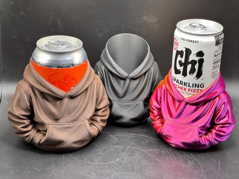 May include: Three hoodie-shaped can holders in brown, gray, and pink. The brown holder has a can with an orange label, the gray holder is empty, and the pink holder has a can with the text "CHI FOREST" and "SPARKLING LYCHEE FIZZY".