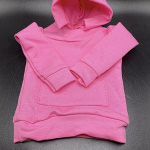 May include: A bright pink hooded sweatshirt with long sleeves and a kangaroo pocket. The sweatshirt is made of a soft, knit fabric and features a drawstring hood and ribbed cuffs and hem.