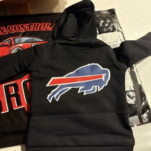 May include: Black hooded sweatshirt featuring the Buffalo Bills logo on the back. The logo is a blue buffalo with a red stripe and white outline. The sweatshirt has a drawstring hood and long sleeves.