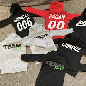 May include: A collection of children's hoodies in various colors, including black, red, and gray. Each hoodie features a hood and long sleeves. Some hoodies have text such as "SANPIETRO 006", "PAGAN 00", "LAWRENCE", "TEAM TRC", and the Nike logo.