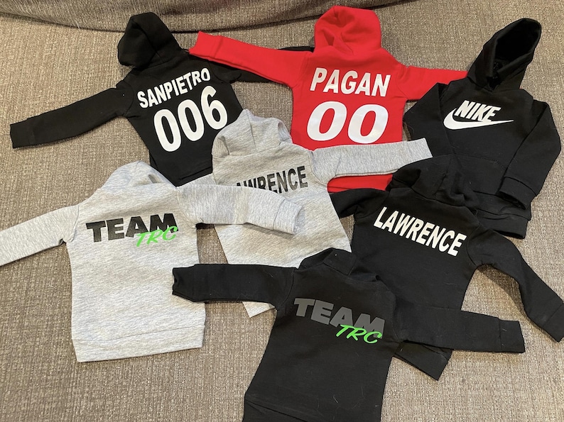 May include: A collection of children's hoodies in various colors, including black, red, and gray. Each hoodie features a hood and long sleeves. Some hoodies have text such as "SANPIETRO 006", "PAGAN 00", "LAWRENCE", "TEAM TRC", and the Nike logo.