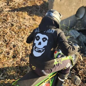 May include: A person wearing a black hoodie with a white skull graphic and the word "Misfits" in white lettering on the back, riding a green and black motorcycle. The person is wearing a helmet and protective gear.
