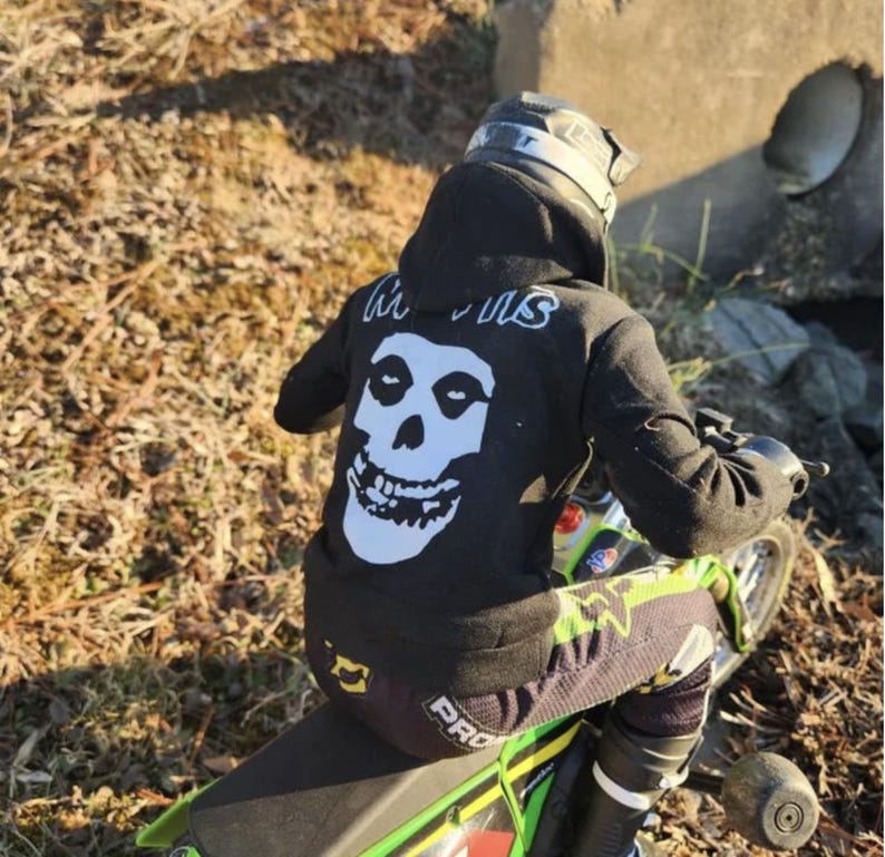 May include: A person wearing a black hoodie with a white skull graphic and the word "Misfits" in white lettering on the back, riding a green and black motorcycle. The person is wearing a helmet and protective gear.