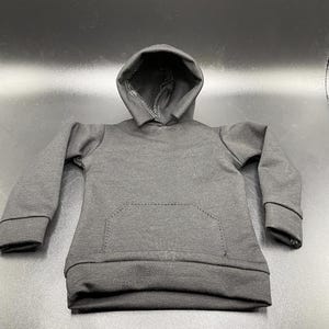 May include: A small, black hooded sweatshirt with a kangaroo pocket. The miniature hoodie is made of a soft fabric and features a drawstring hood and ribbed cuffs. The garment is designed for a doll or action figure.