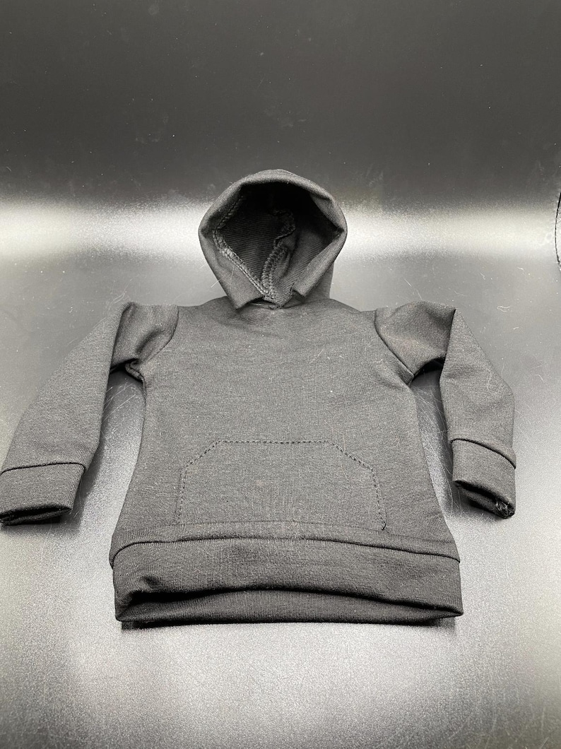 May include: A small, black hooded sweatshirt with a kangaroo pocket. The miniature hoodie is made of a soft fabric and features a drawstring hood and ribbed cuffs. The garment is designed for a doll or action figure.