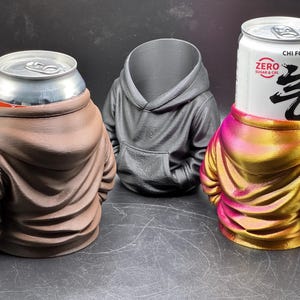 May include: Three beverage can holders shaped like hoodies. One is brown, one is black, and the third is a gradient of pink and gold. The can holders are designed to hold standard-sized beverage cans.