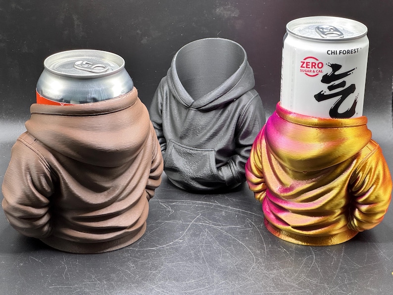May include: Three beverage can holders shaped like hoodies. One is brown, one is black, and the third is a gradient of pink and gold. The can holders are designed to hold standard-sized beverage cans.