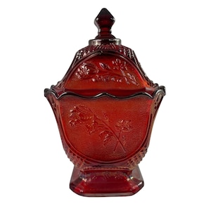May include: A ruby red, lidded candy dish with a textured surface and floral relief design. The dish has a pedestal base and a pointed finial on the lid. The candy dish is a vintage piece.