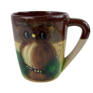 Puede incluir: Una taza de cerámica con exterior marrón y asa. La taza presenta un diseño de búho pintado a mano en tonos marrones, beige y verdes. Los ojos del búho son grandes y negros con reflejos amarillos. La taza es adecuada para bebidas calientes.