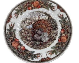 Può includere: Un piatto in ceramica bianca con bordo smerlato caratterizzato da un'illustrazione centrale di un tacchino in un paesaggio boschivo con una capanna. Il bordo è decorato con un colorato motivo autunnale di zucche, mais, uva e foglie.