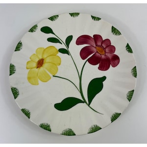 May include: White ceramic plate with a hand-painted floral design. The plate features a yellow and a burgundy flower with green leaves and stems. The rim is decorated with green dots. A decorative piece for serving or display.
