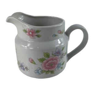 Op de afbeelding: Een witte keramische kan met een grijze handgreep en tuit. De kan is versierd met roze, paarse en blauwe bloemmotieven. Een klassiek ontwerp voor het serveren van dranken.