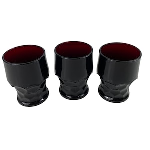 Puede incluir: Tres vasos con un exterior negro profundo y un interior rojo oscuro en contraste. Cada vaso tiene un borde superior liso y un patrón geométrico facetado y texturizado alrededor de la base, creando un contraste visual distintivo.