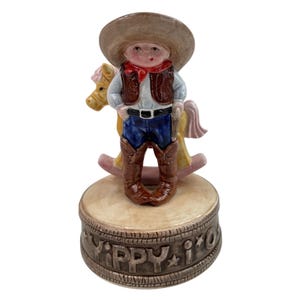 Puede incluir: Figura de vaquero de cerámica con sombrero beige, pañuelo rojo y chaleco marrón. La base está decorada con las palabras "YIPPY I O". Un caballito balancín amarillo está detrás del vaquero.