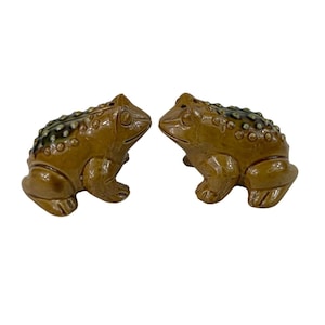 May include: A pair of ceramic frog figurines, likely salt and pepper shakers, in a glossy brown glaze. The frogs have textured backs with dark green and brown accents. The figurines are posed facing each other.