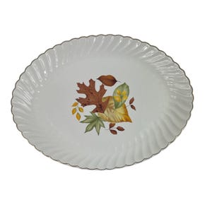 Puede incluir: Una bandeja de servir ovalada de color blanco con un borde festoneado y acanalado, y un borde metálico plateado. El centro de la bandeja presenta una ilustración decorativa de varias hojas de otoño en tonos marrón, amarillo, verde y naranja.