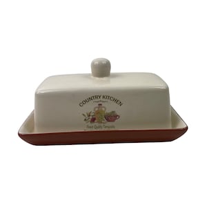 May include: A cream-colored ceramic butter dish with a rectangular base and a matching lid featuring a knob handle. The front displays the text "COUNTRY KITCHEN" above a graphic of a bottle and bowl, with "Finest Quality Terracotta" below.