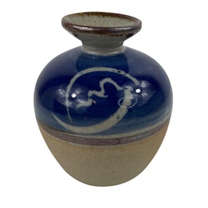 May include: A small, round ceramic vase with a dark blue glazed top and a tan base. The blue section features a white, abstract design. The rim is brown, and the vase has a bulbous shape, perfect for displaying small flowers or as a decorative piece.