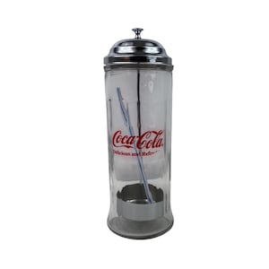 Puede incluir: Un dispensador de pajitas Coca-Cola de cristal transparente con tapa de cromo pulido y una varilla metálica vertical en el interior. El frente muestra el logotipo rojo de Coca-Cola y el texto Delicious and Refreshing impreso.