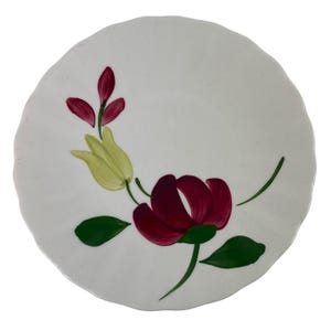 May include: A white ceramic plate with a scalloped edge featuring a hand-painted floral design. The pattern includes a large dark red flower, a pale yellow tulip-like bud, smaller red buds, and green leaves painted with visible brushstrokes.